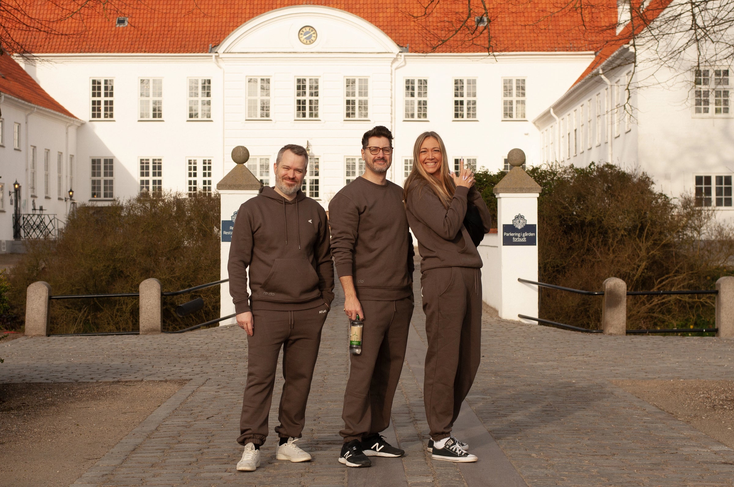 re do x Atlab collab. Smilende team står samlet iført ens firmatøj med tryk fra re do® og tøj med logo fra re do® foran klassisk bygning.